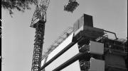 A large crane sits in front of a partially completed brutalist style building. 