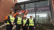 Four men in high ves vests and hard hats pose for a photo in front of the foyer of the WA Museum.  A small construction machine with a crane arm is behind them ready to smash the glass foyer. 