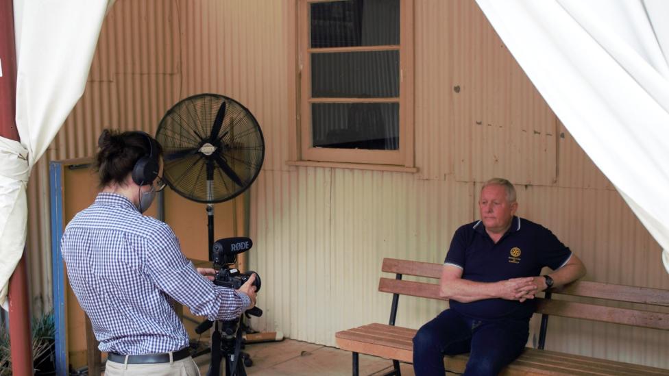 A person sits on a bench in front of a camera, which is being operated by another person wearing a light-blue long sleeved shirt