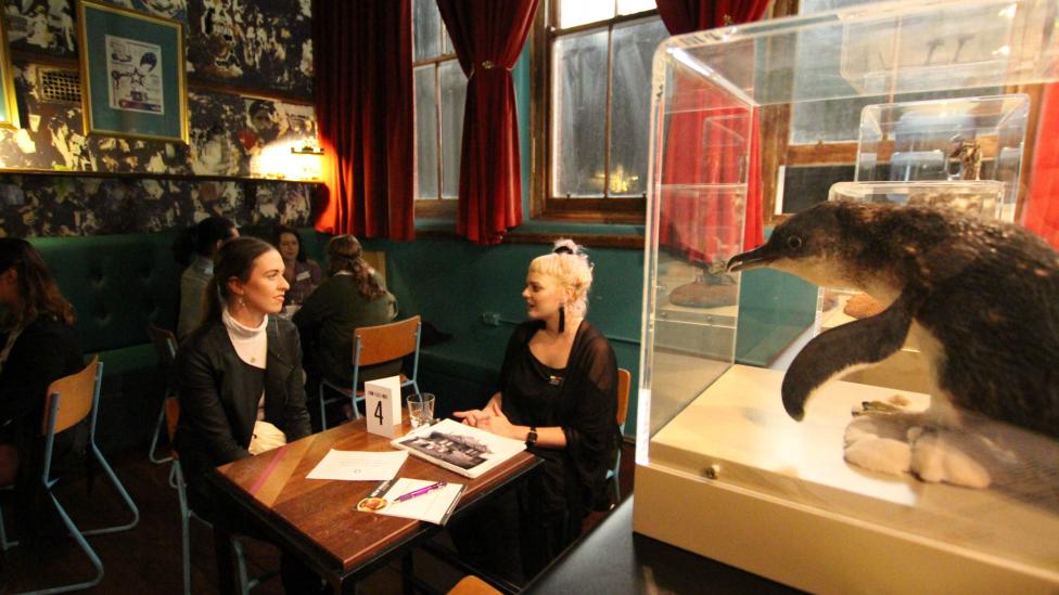 "Two young women talking at a table, taxidermied penguin in a perspex case in the foreground."