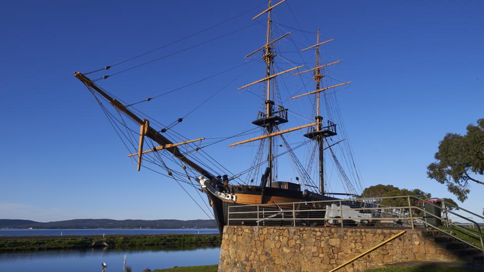 A Brig is moored near a stone wall beneath a clear blue sky