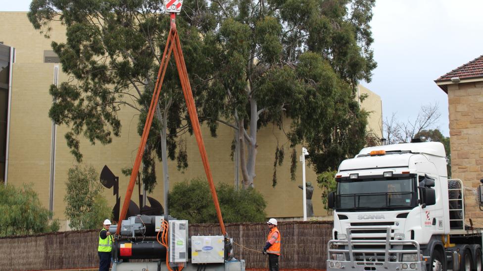 Two workers holding the straps around a large chiller.  The chiller is about to be craned onto the roof of the Art Gallery of WA 