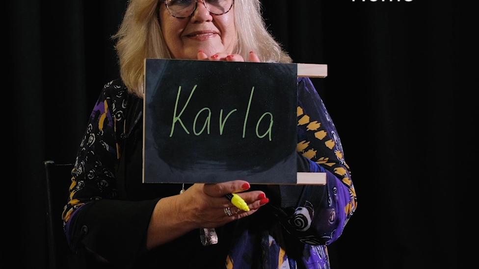 "A woman holds up a small chalkboard that reads 'Karla'. Text reads 'Home'."