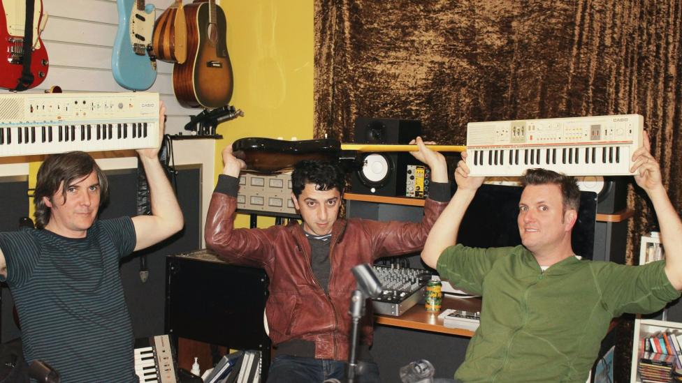 Three men sit in a recording studio holding casio keyboards over their heads.