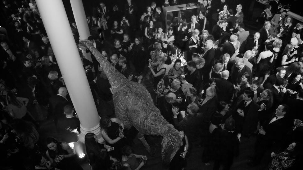 "A high shot of a crowd partying in Hackett Hall. A dinosaur is moving among them."