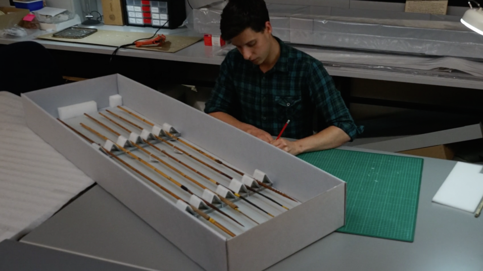 Person sitting at a desk writing, behind a box filled with arrows. 