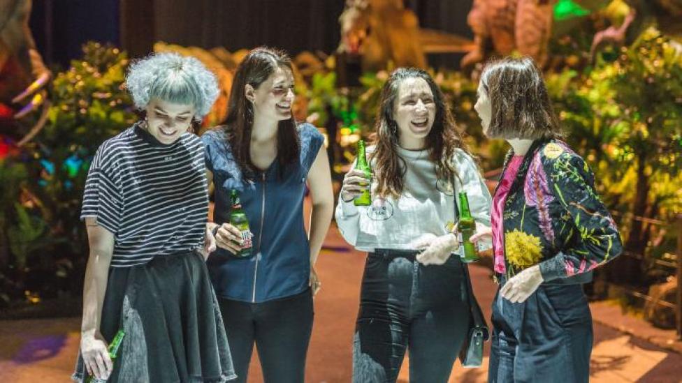 "A group of people drinking and standing around an animatronic dinosaur."