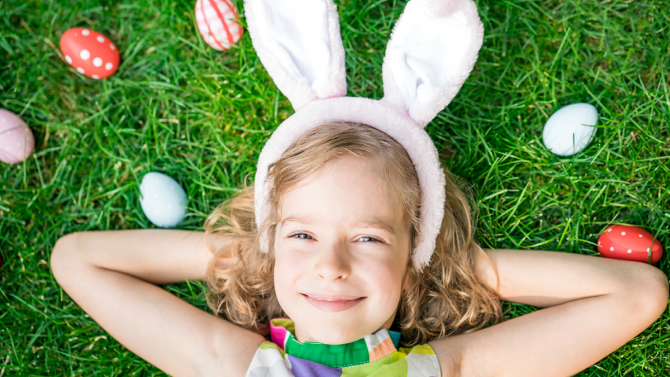 "A young girl with bunny ears on, lying down surrounded by Easter eggs."