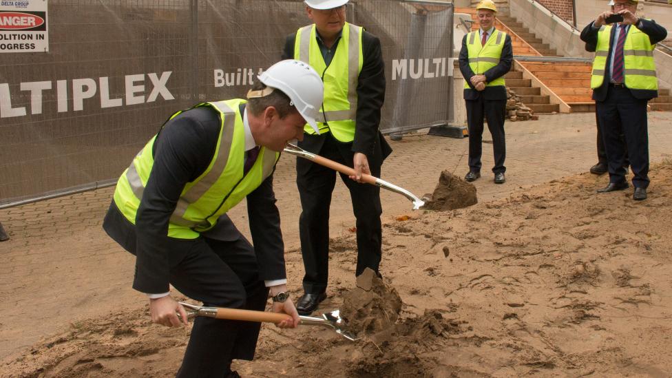 Premier McGowan bending with a ceremonial shovel and Minister Templeman standing to his left.  They are turning sod by digging the soil at the site of the New Museum.  Both are wearing fluro yellow vests and hard hats. 