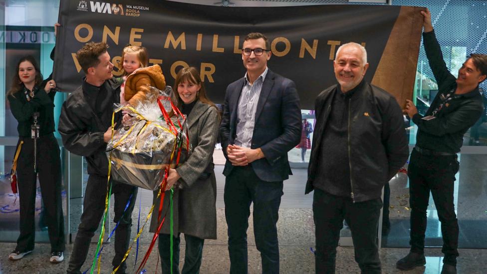 Four adults and a toddler stand in front of a banner that says "One Millionth Visitor" held by two Museum staff members.