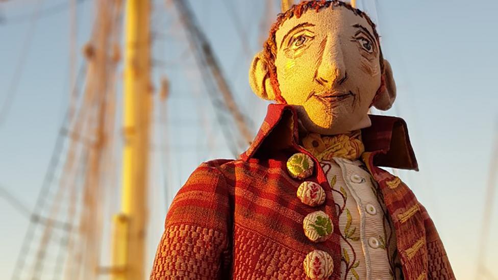 A handcrafted puppet of an 18th Century botanist with a boat in the background.