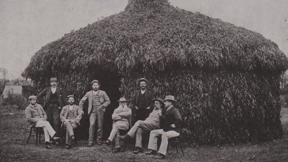 A crowd of people in front a large grass hut