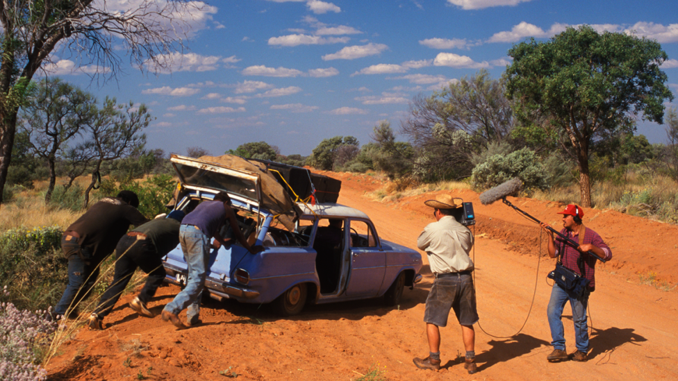 Three mechanics push a blue car under repair in the bush while two people film them for the Bush Mechanics TV series.