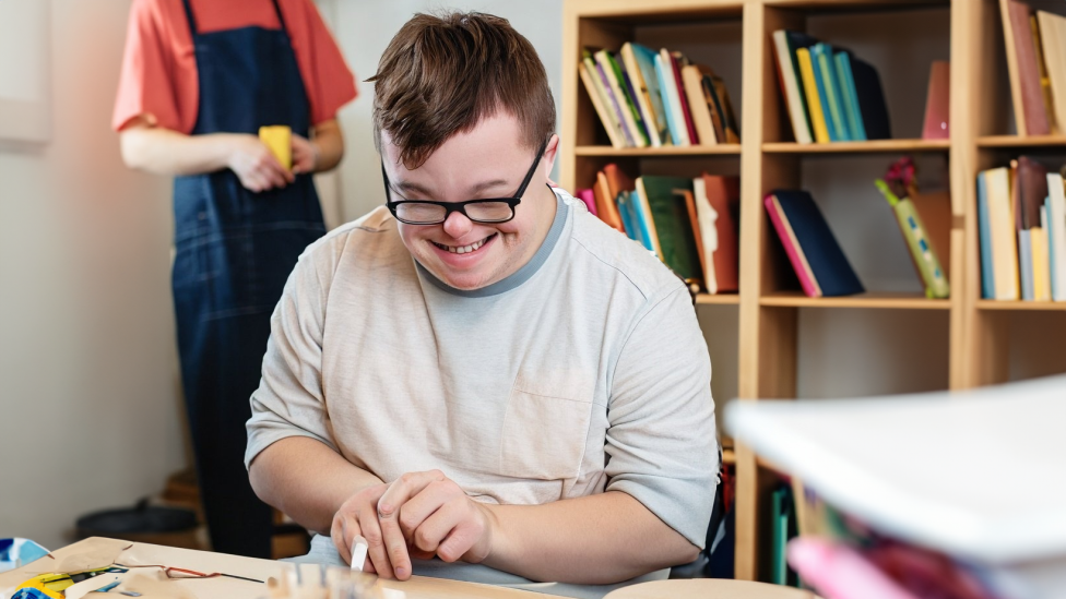 A young man with downs syndrome smiles down at a craft project they're making with string and popsicle sticks