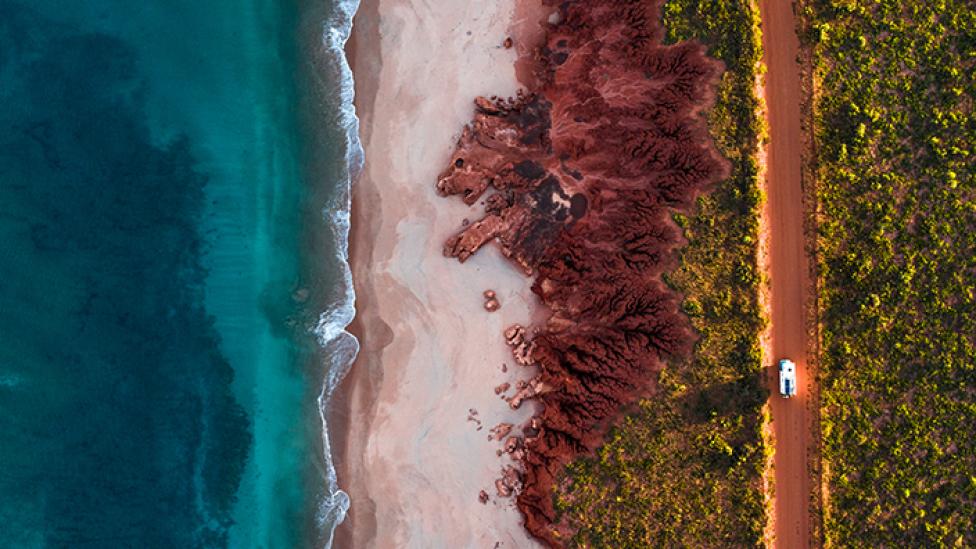 Ocean, red dirt and car driving along a dusty road - aerial shot.