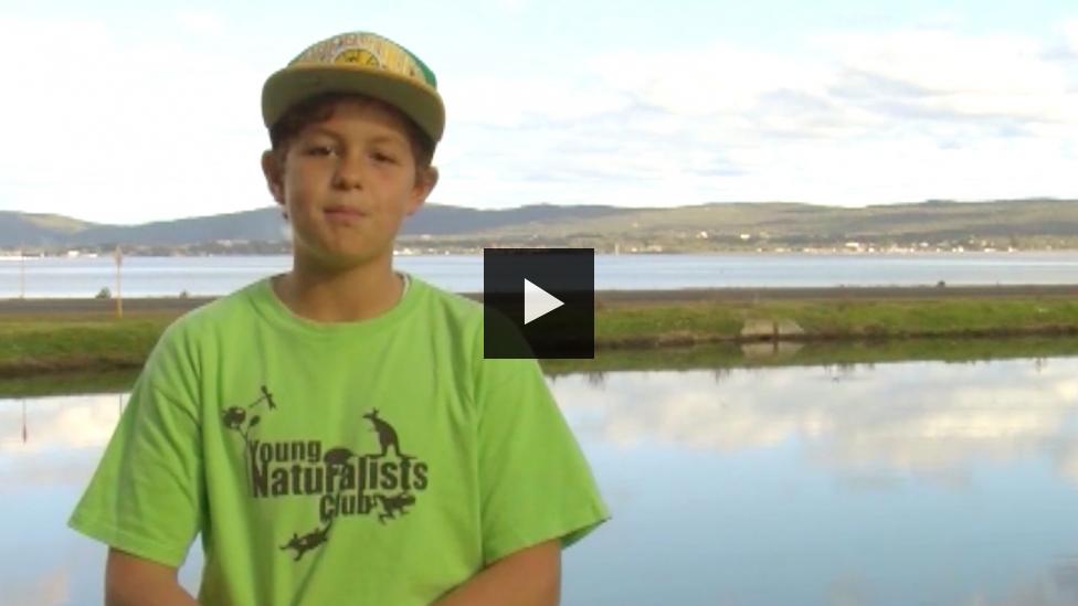 Boy in Young Naturalists Club T-Shirt stand in front of a lake and hills
